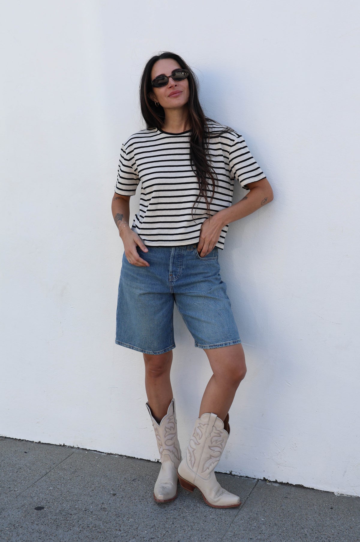 Woman wearing a striped t-shirt with jean shorts and boots against a white wall