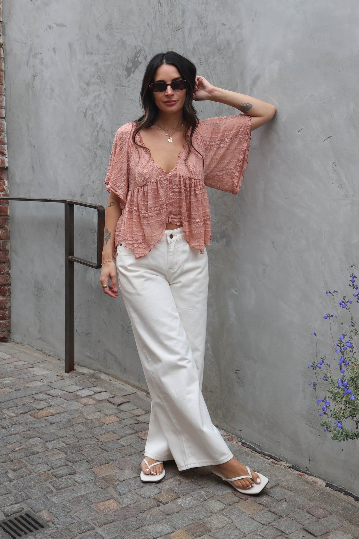 Woman wearing white jeans and a blush lace blouse against a brick wall