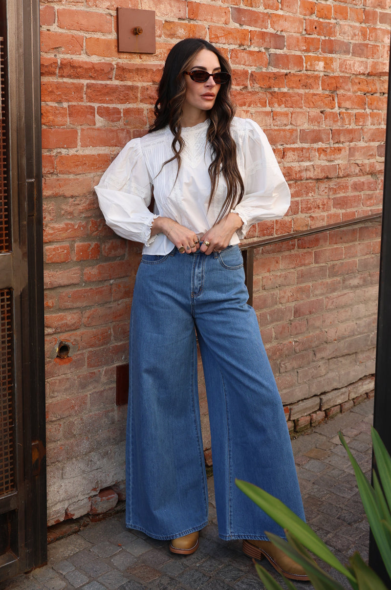Woman in a white blouse and blue jeans standing against a brick wall.