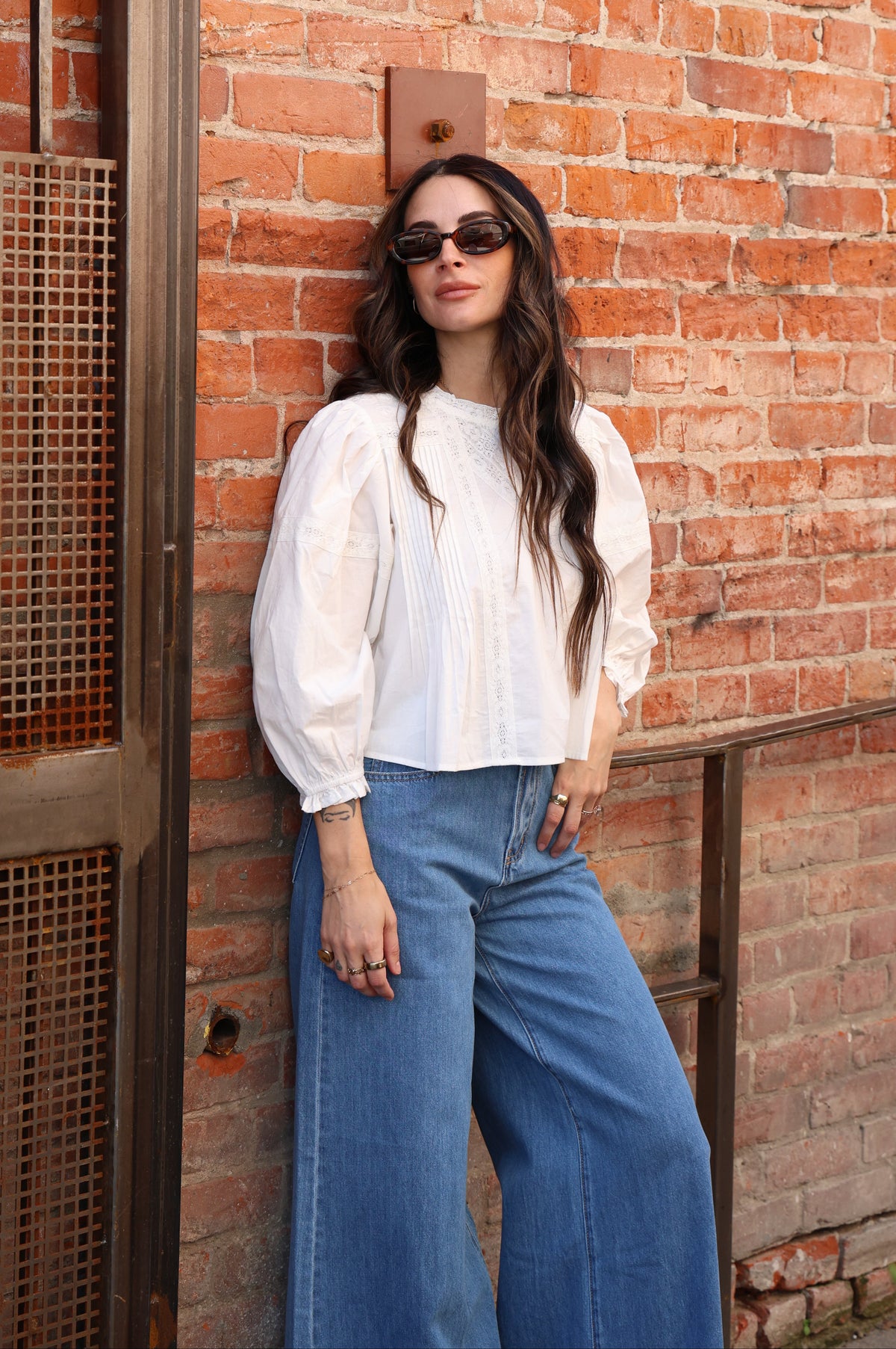 Woman in a white blouse and blue jeans standing against a brick wall.