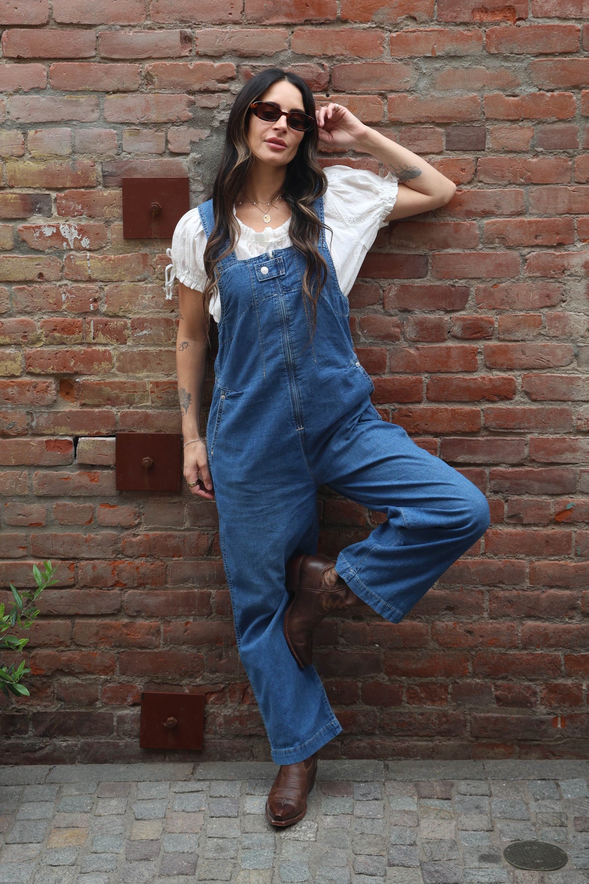 Woman wearing a pair of overalls with a white blouse against a brick wall