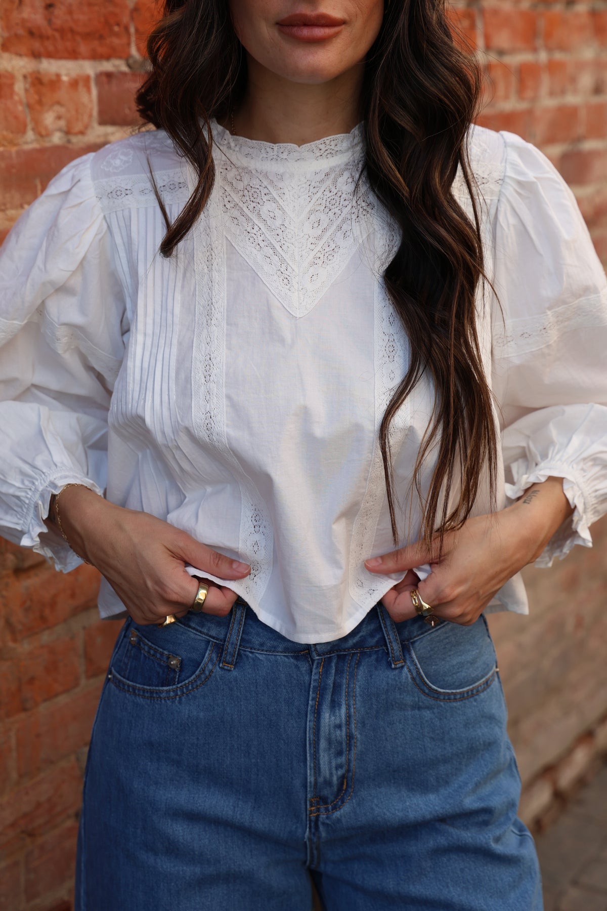 Person wearing a white blouse with lace details and blue jeans against a brick wall.