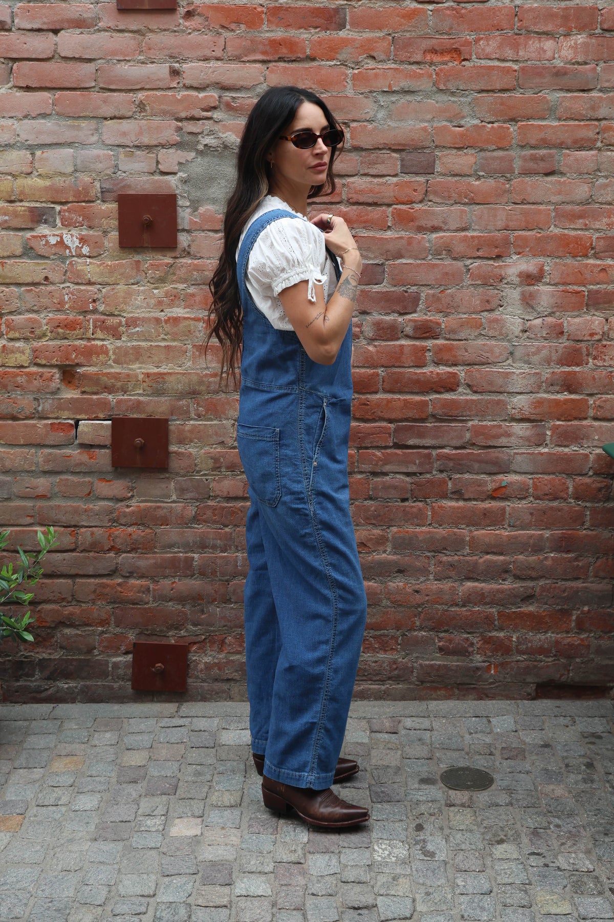 Woman wearing a pair of overalls with a white blouse against a brick wall