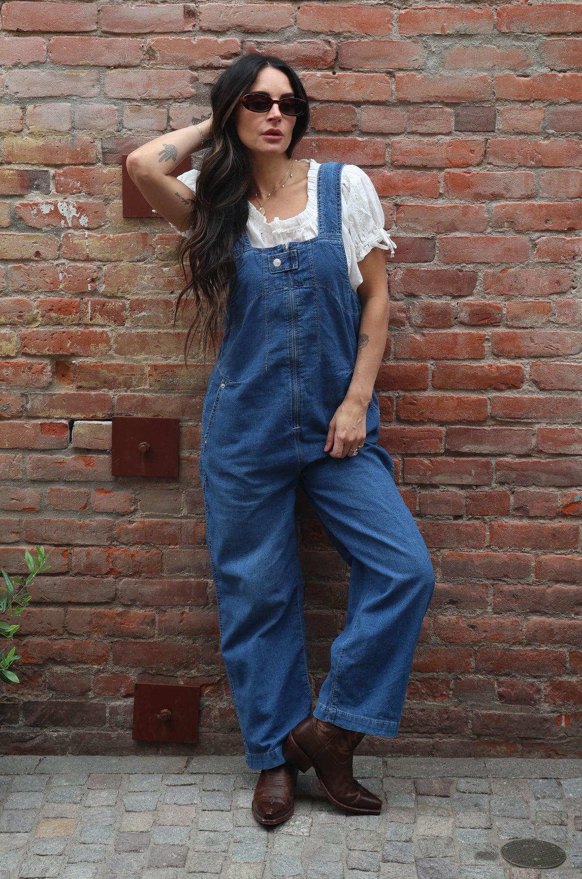 Woman wearing a pair of overalls with a white blouse against a brick wall