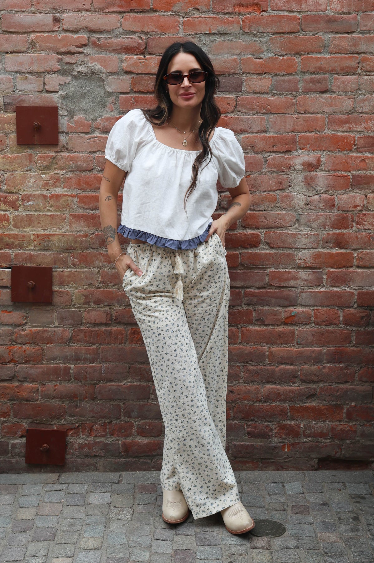 Woman wearing a white blouse with a blue ruffle trim and flower pants against a brick wall
