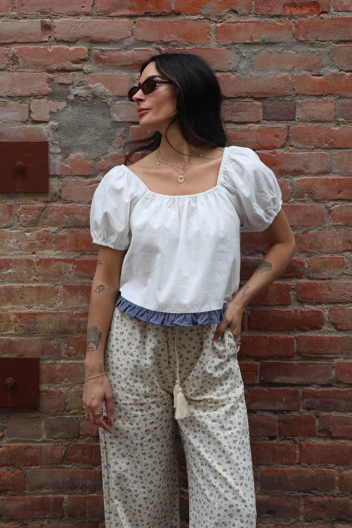 Woman wearing a white blouse with a blue ruffle trim and flower pants against a brick wall