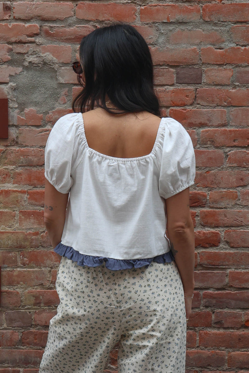 Woman wearing a white blouse with a blue ruffle trim and flower pants against a brick wall