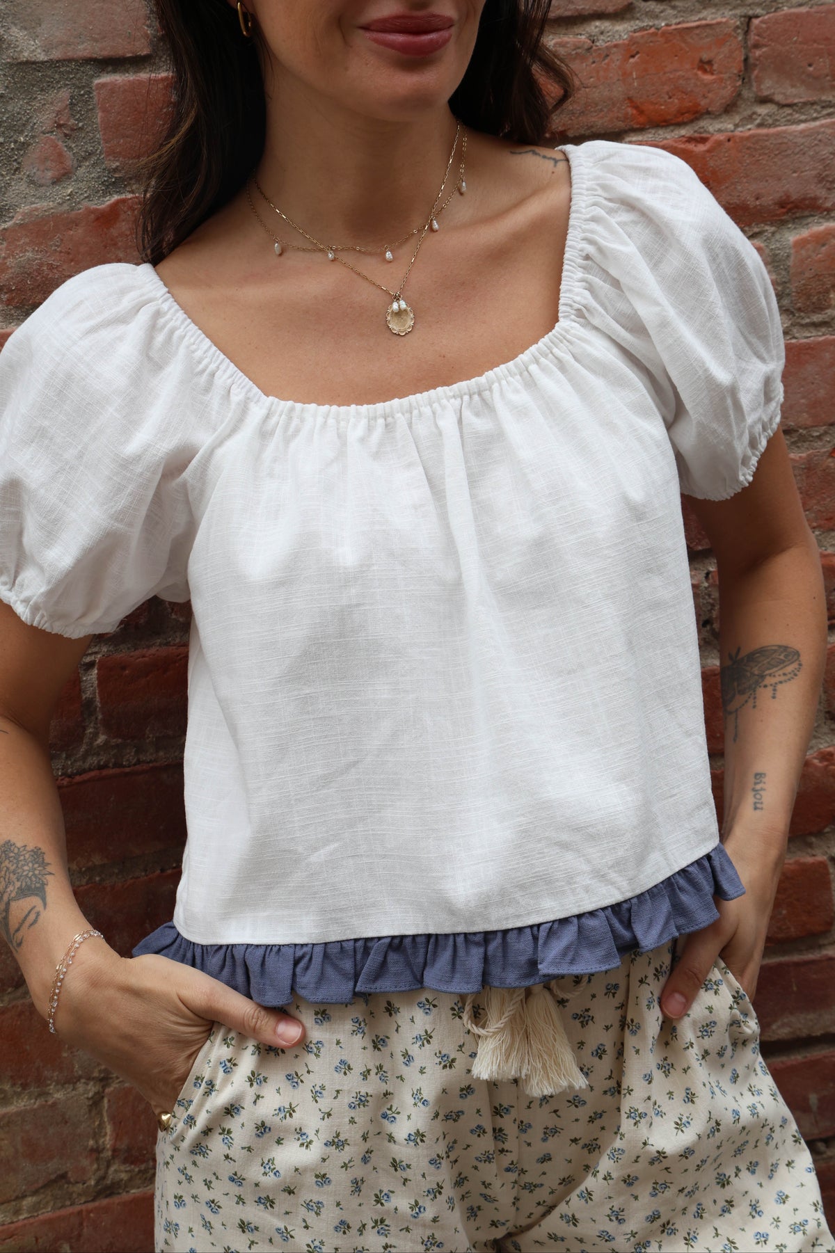 Woman wearing a white blouse with a blue ruffle trim and flower pants against a brick wall
