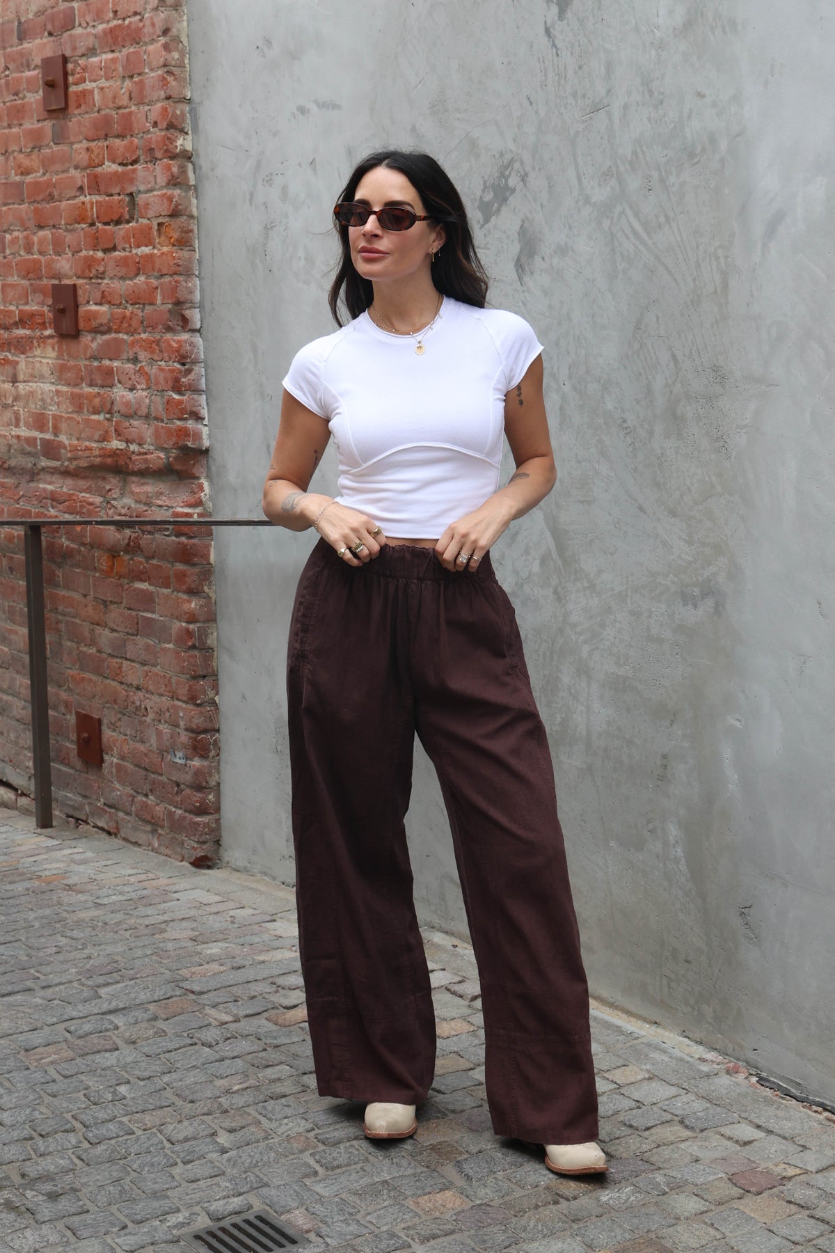 Woman wearing a white top and brown linen pants against a grey background