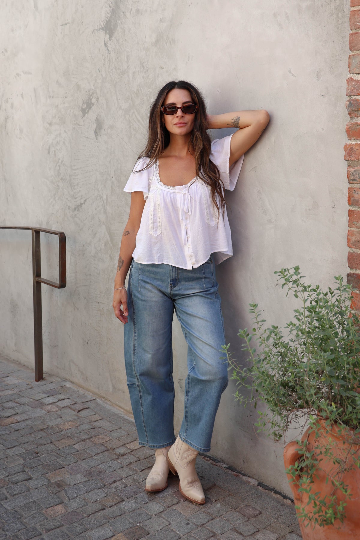 Woman wearing a white blouse with lace details and jeans against a grey wall.