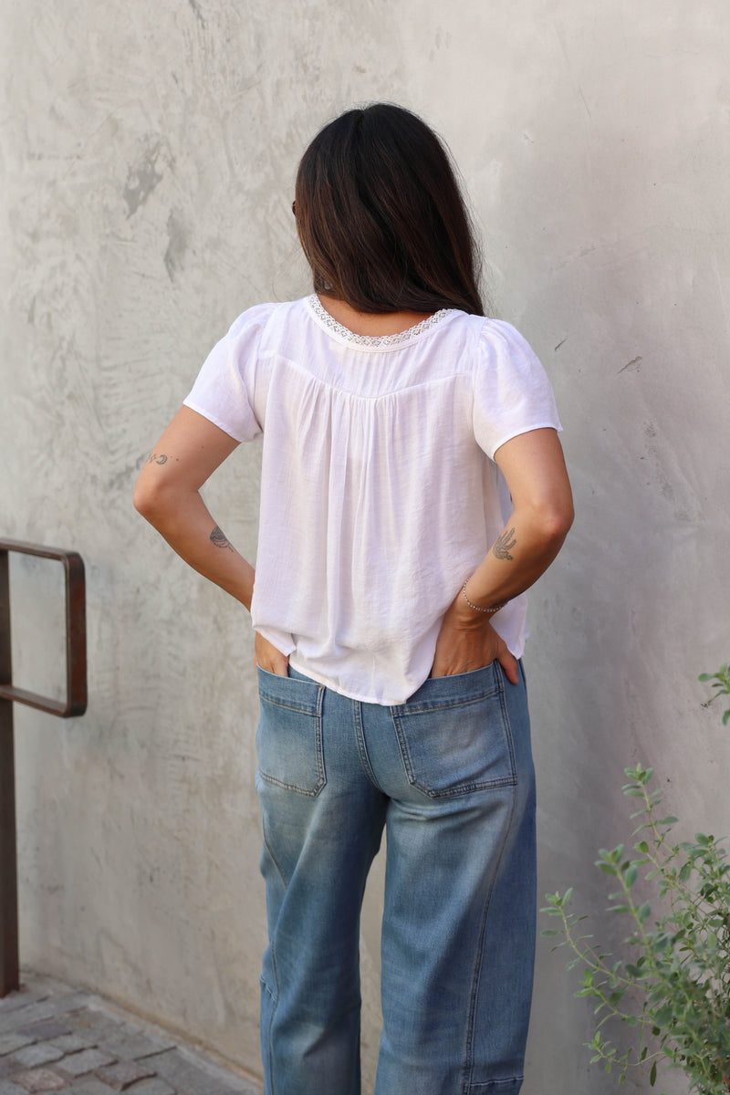 Woman wearing a white blouse with lace details and jeans against a grey wall.