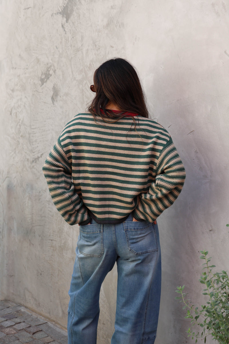 A woman wearing a striped knit sweater with a red crew neck and blue jeans, leaning against a textured gray concrete wall.
