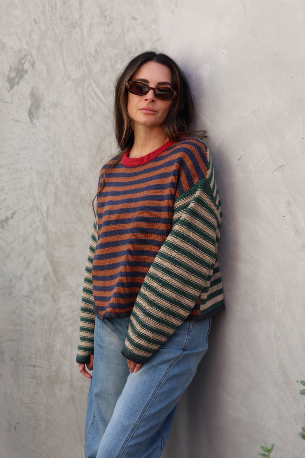 A woman wearing a striped knit sweater with dark green, rust orange, and beige horizontal stripes, paired with light blue jeans and dark sunglasses, leaning against a textured gray concrete wall.