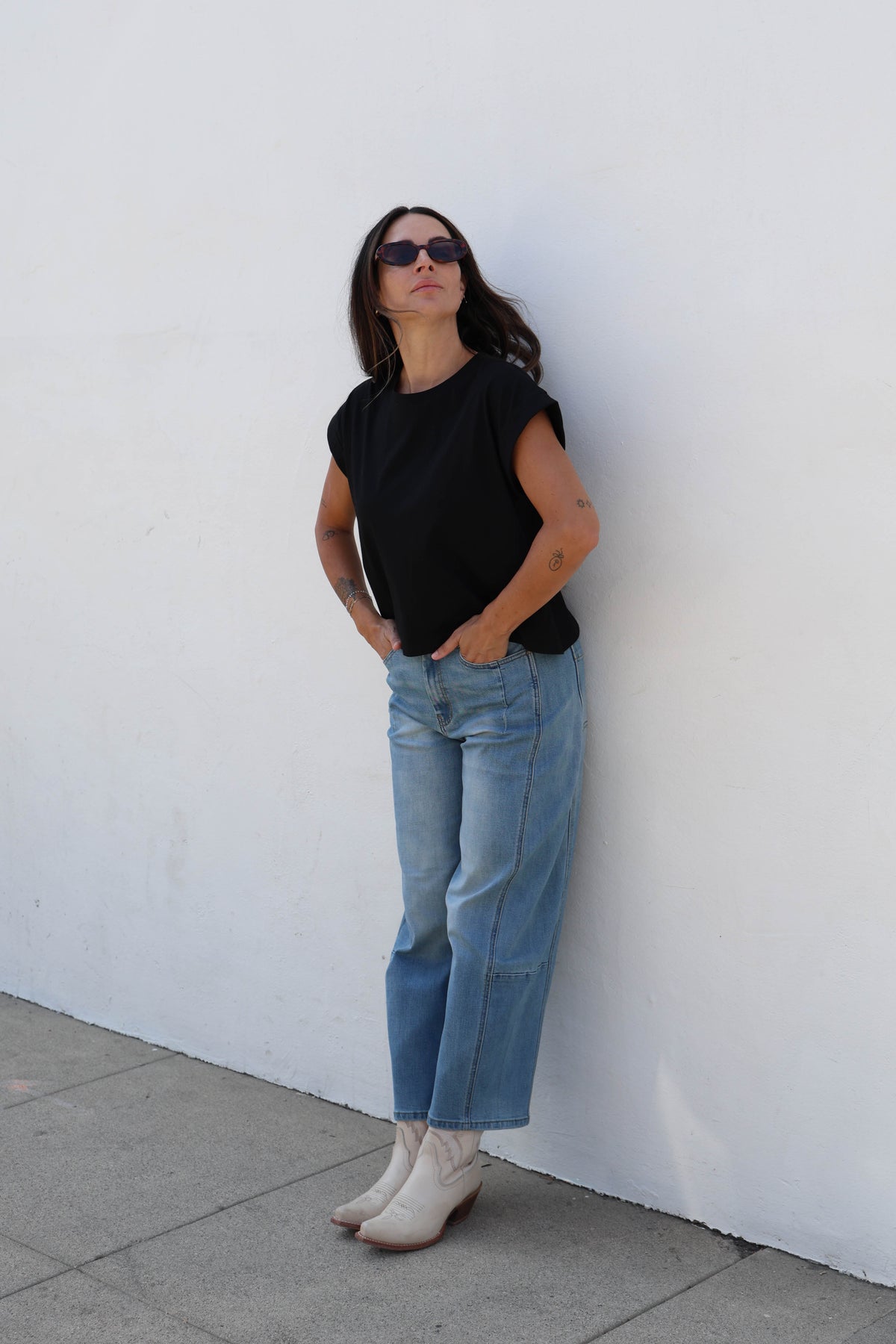 A woman wearing a black short-sleeve top, light blue cropped jeans, and off-white wedge boots stands against a plain white wall.