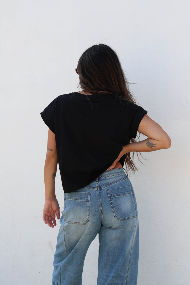 Woman wearing a plain black tee with jeans against a white wall.