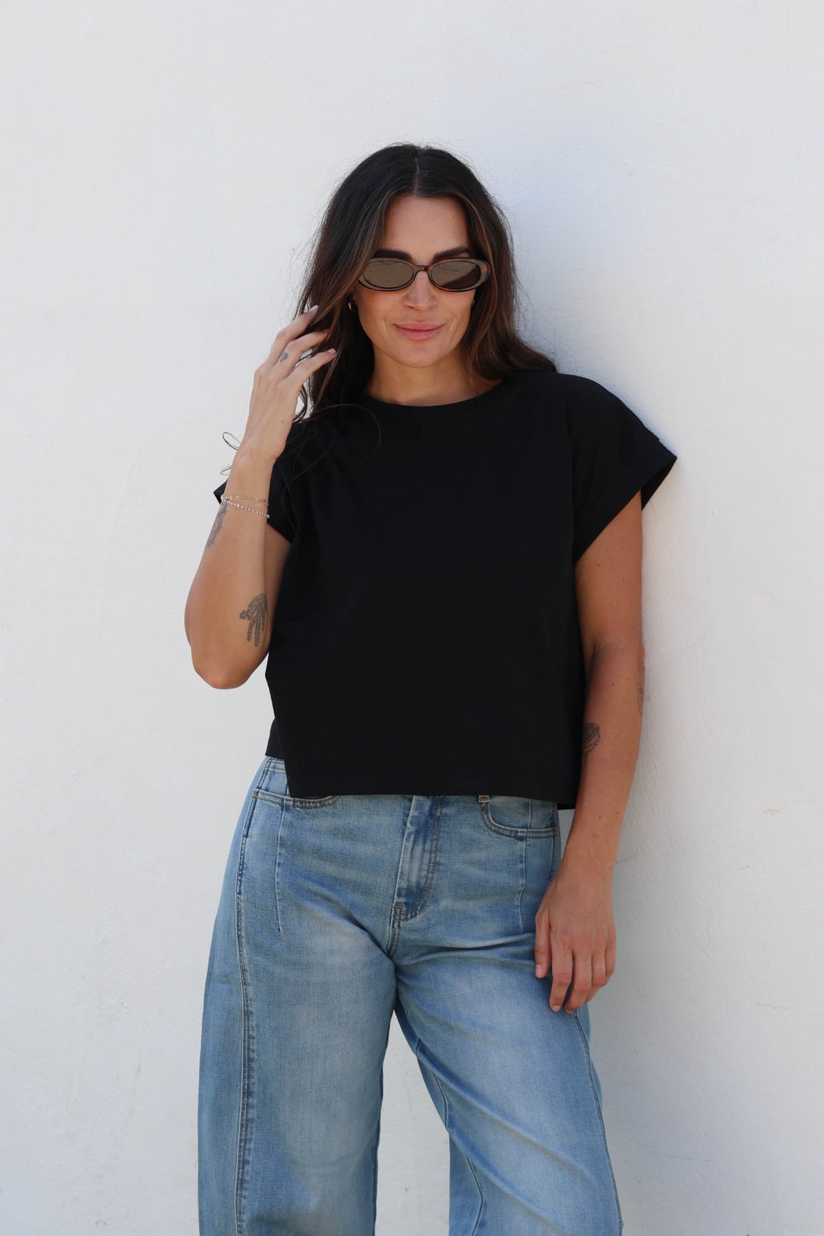 Woman wearing a plain black tee with jeans against a white wall.