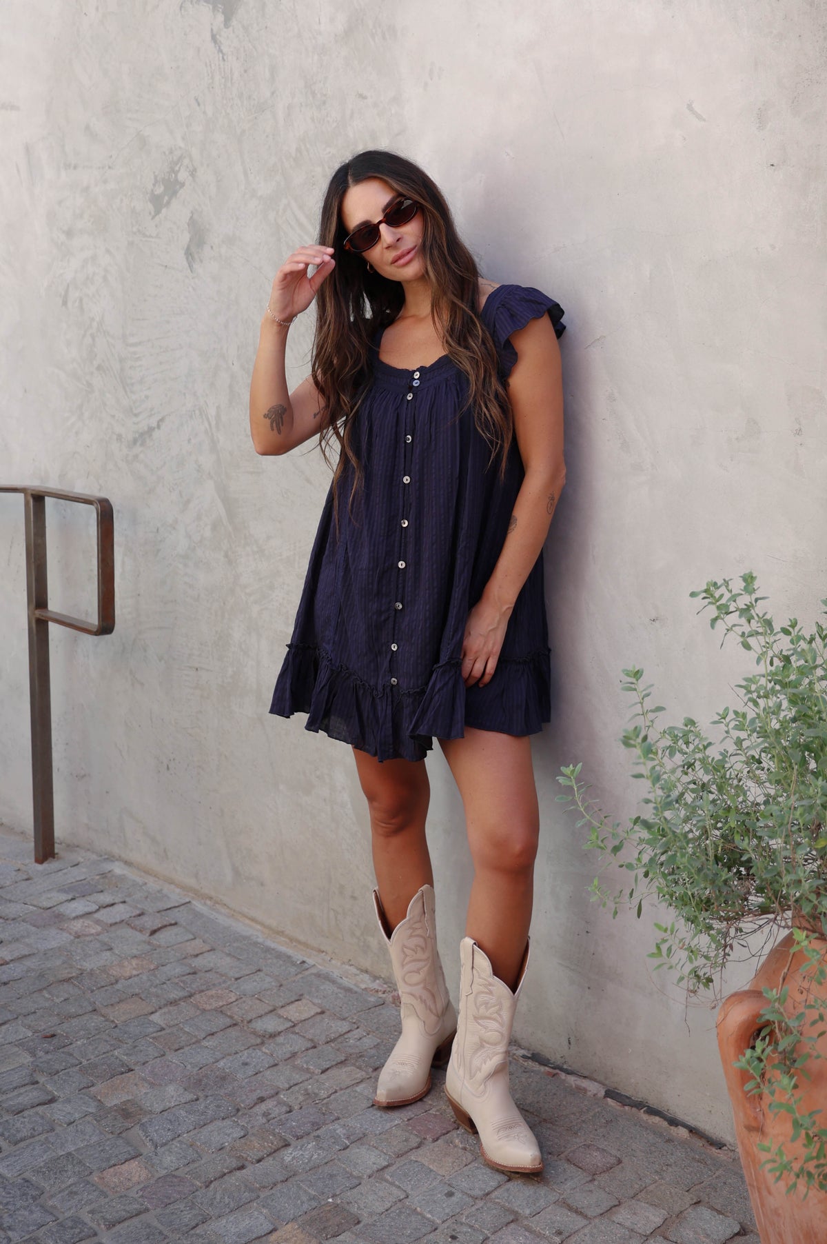 Woman wearing a navy blue dress and cowboy boots against a grey background.