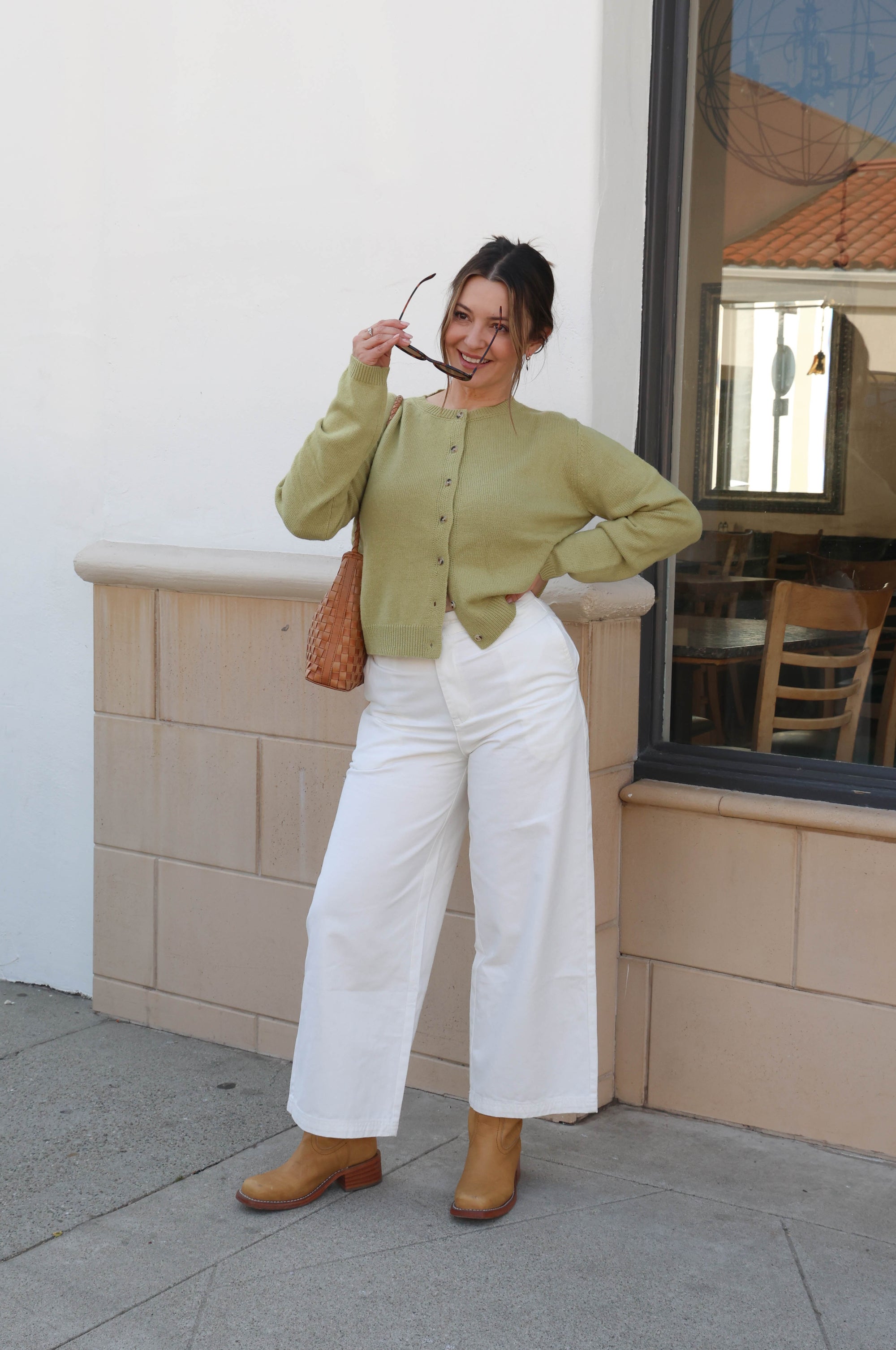 Woman in a green cardigan and white pants standing outside a building.
