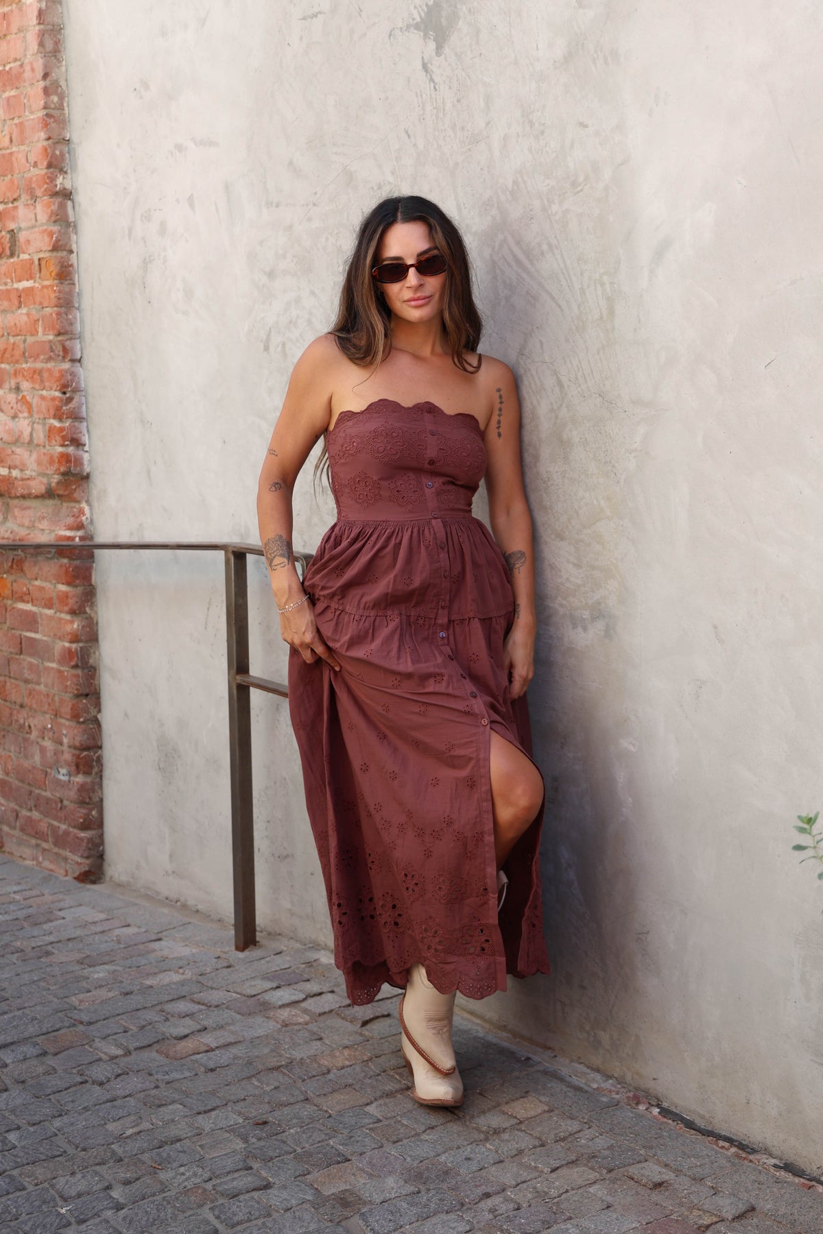 A woman wearing a strapless rust-brown tiered midi dress with eyelet lace details and nude heels, posing against a textured concrete wall.