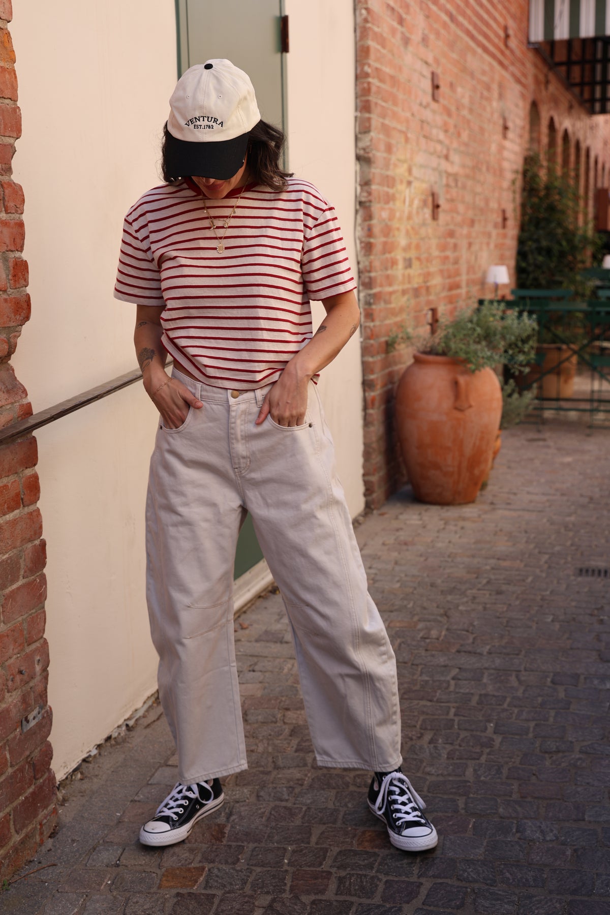 Person wearing a red and white striped shirt, white pants, and black sneakers standing on a brick pavement.