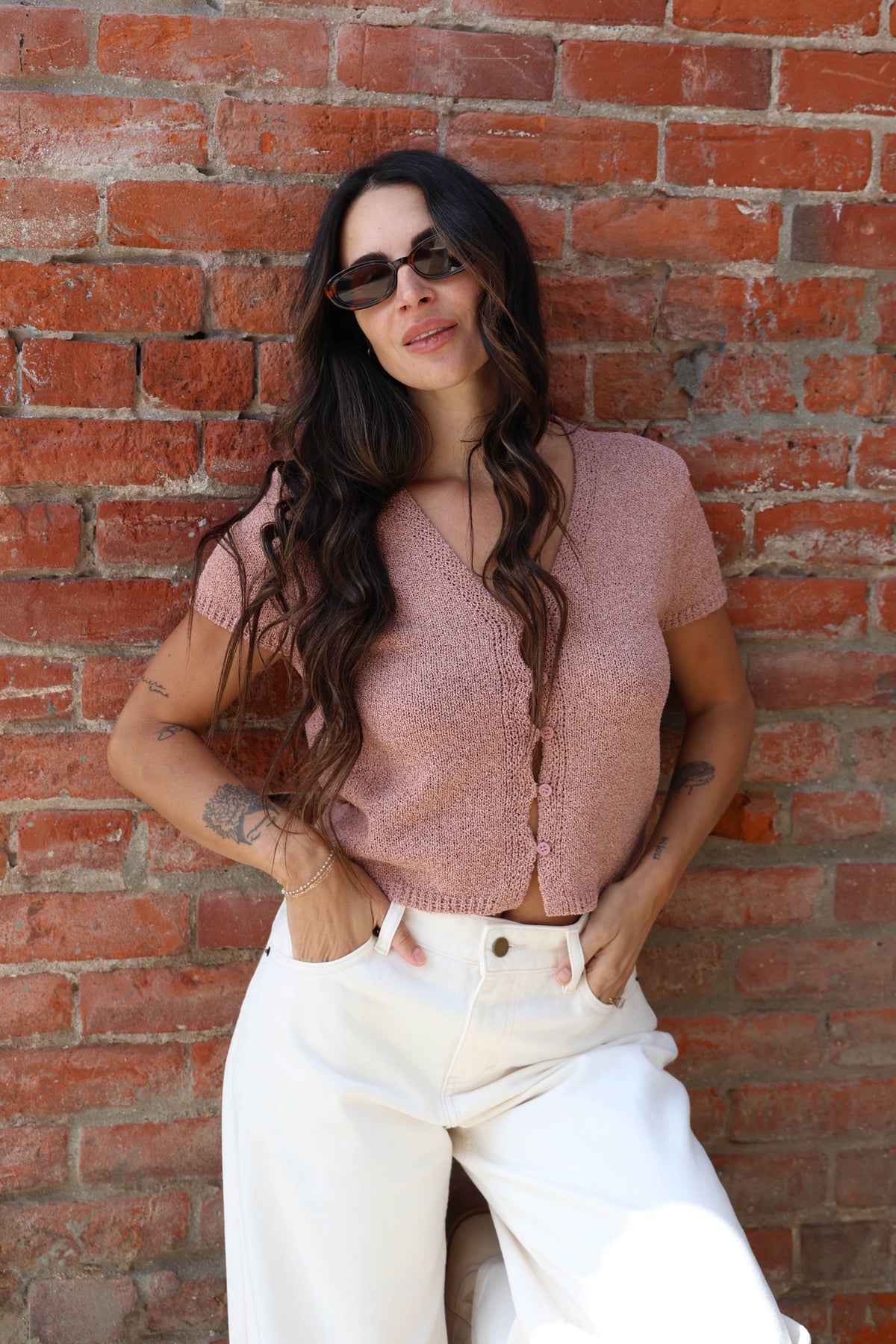 Woman in a pink button up blouse with with jeans against a brick wall