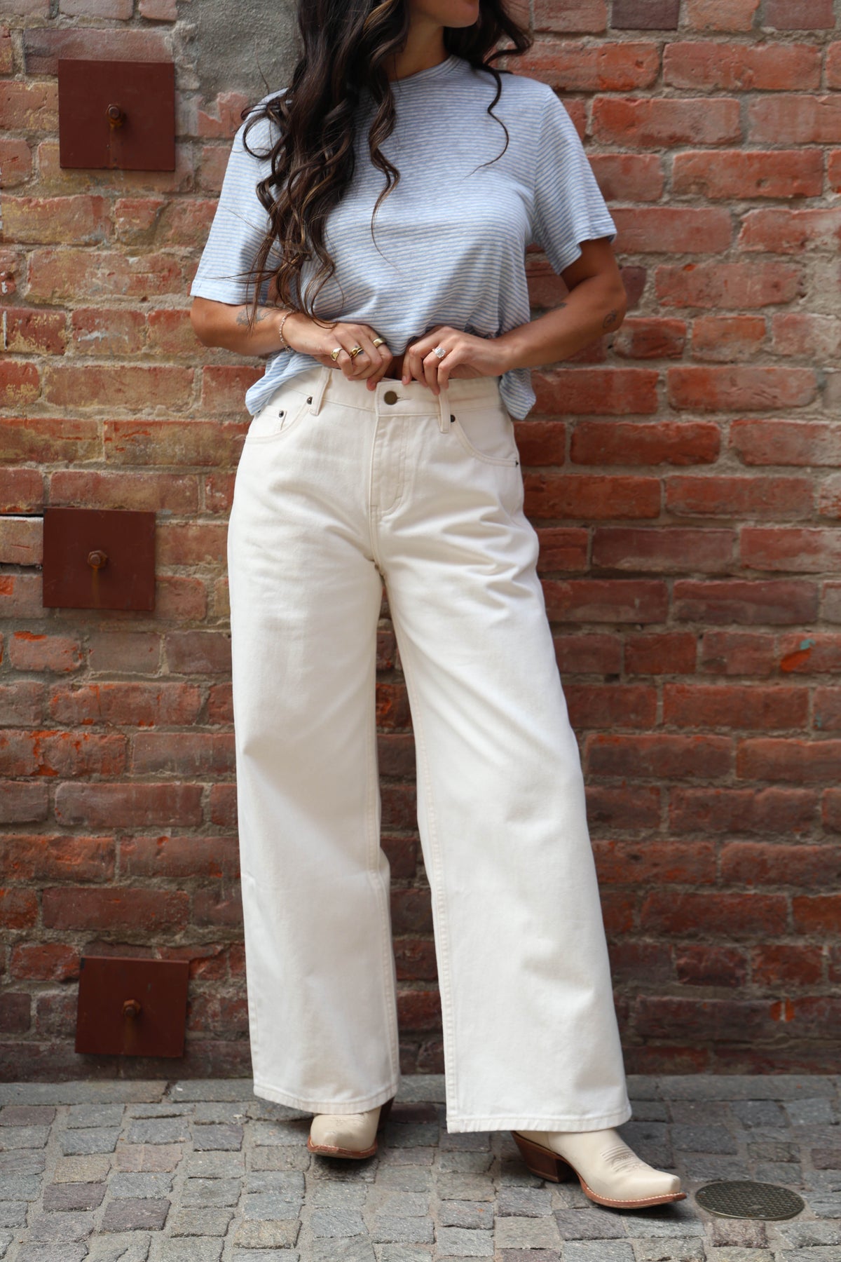 Person wearing a light blue t-shirt and white pants standing against a brick wall.