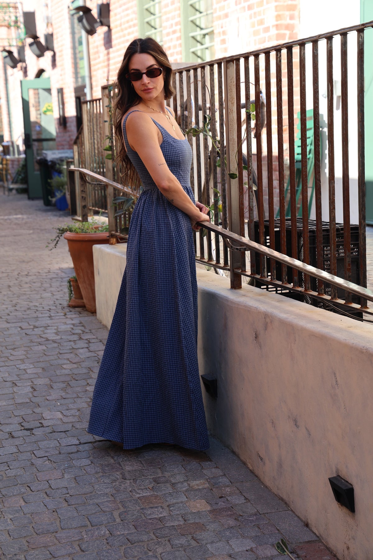 Woman in a blue dress standing on a sidewalk with plants and a railing in the background.