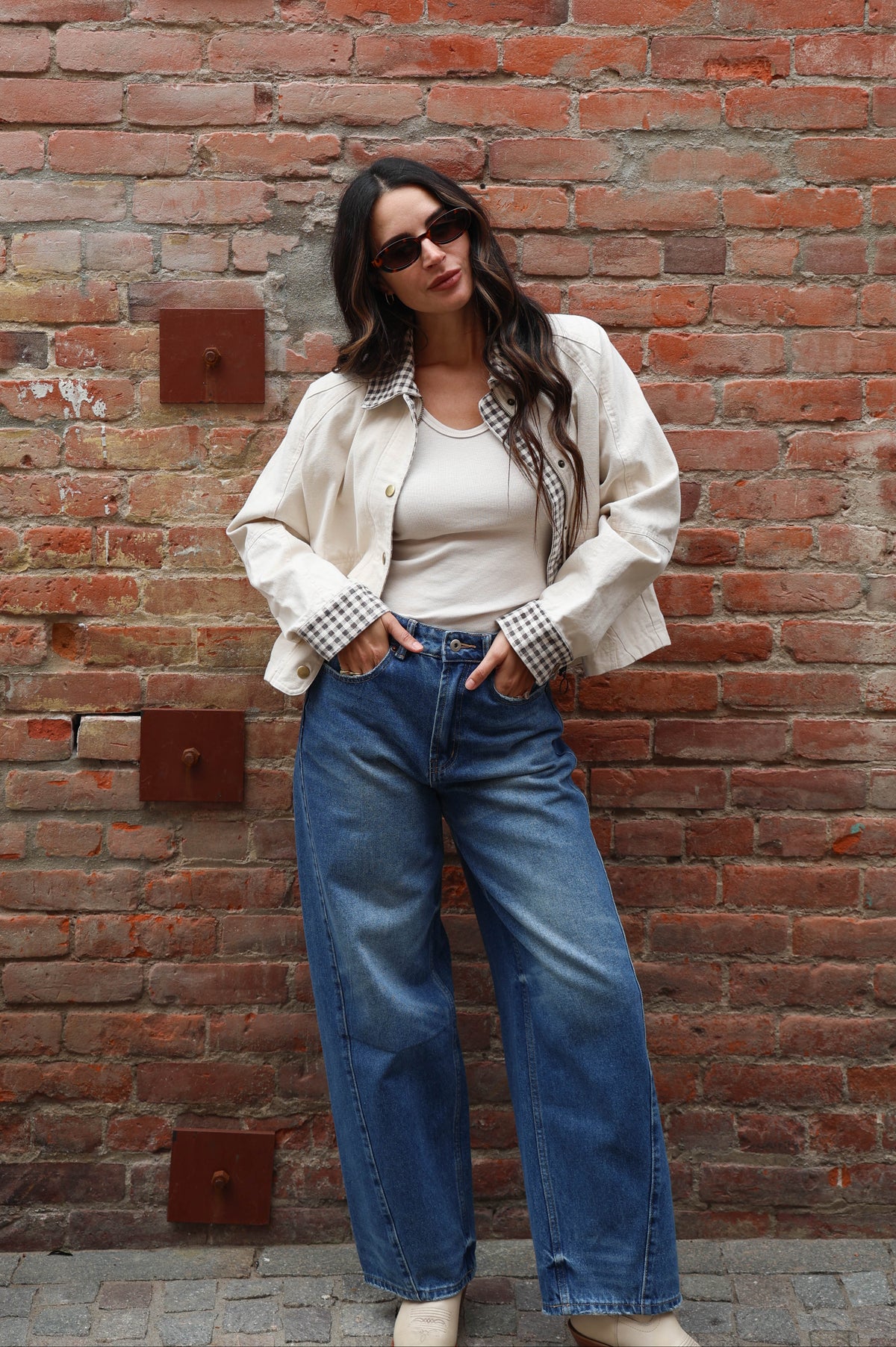 Woman in a beige jacket with gingham details and jeans against a brick wall