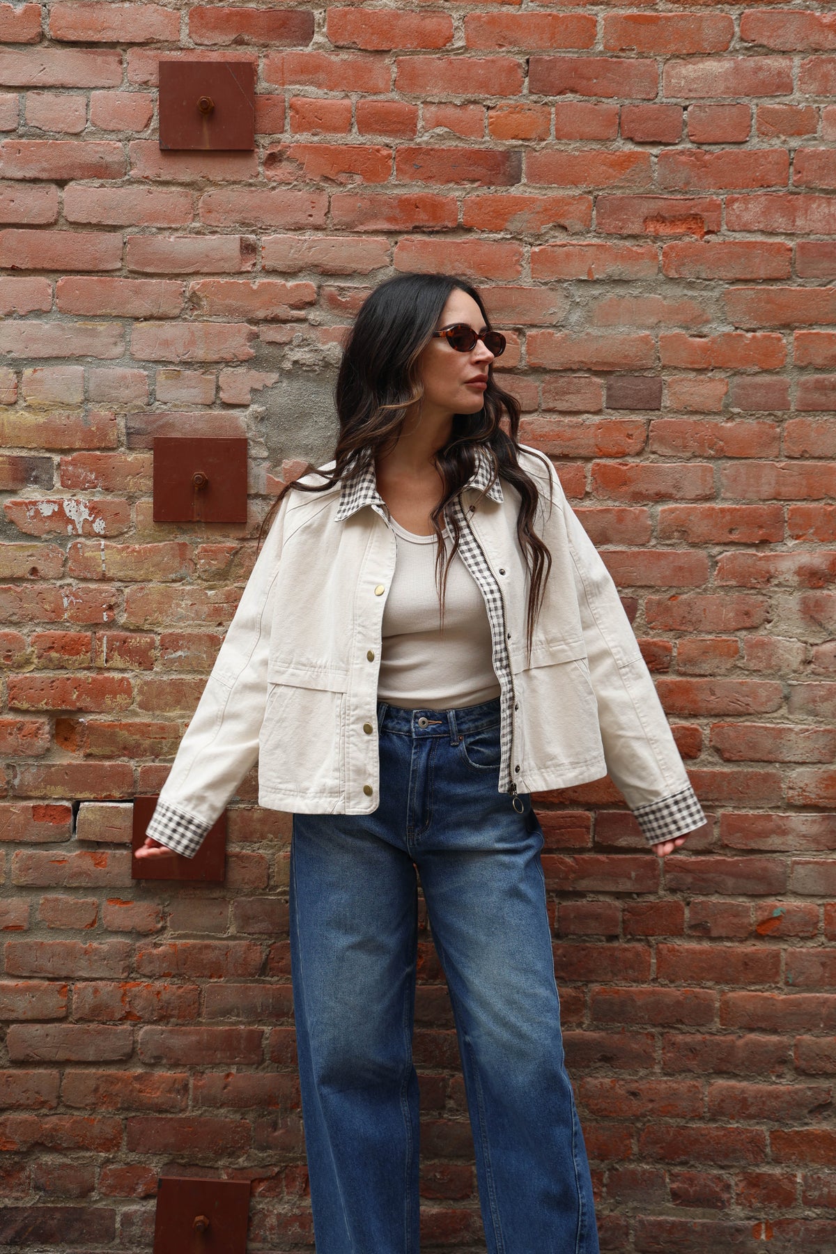 Woman in a beige jacket with gingham details and jeans against a brick wall