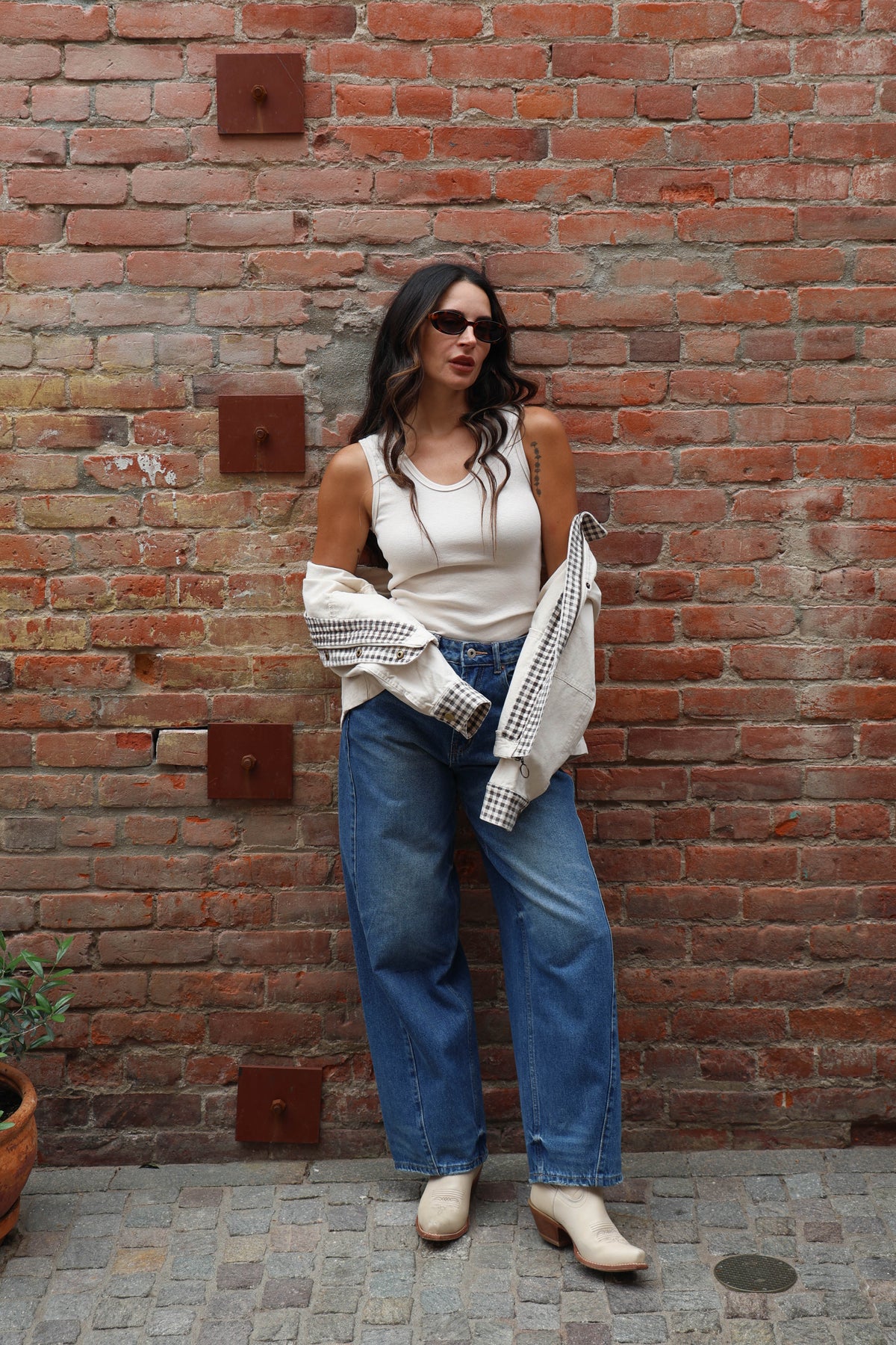 Woman standing against a brick wall wearing a white sleeveless top, blue jeans, and beige boots.
