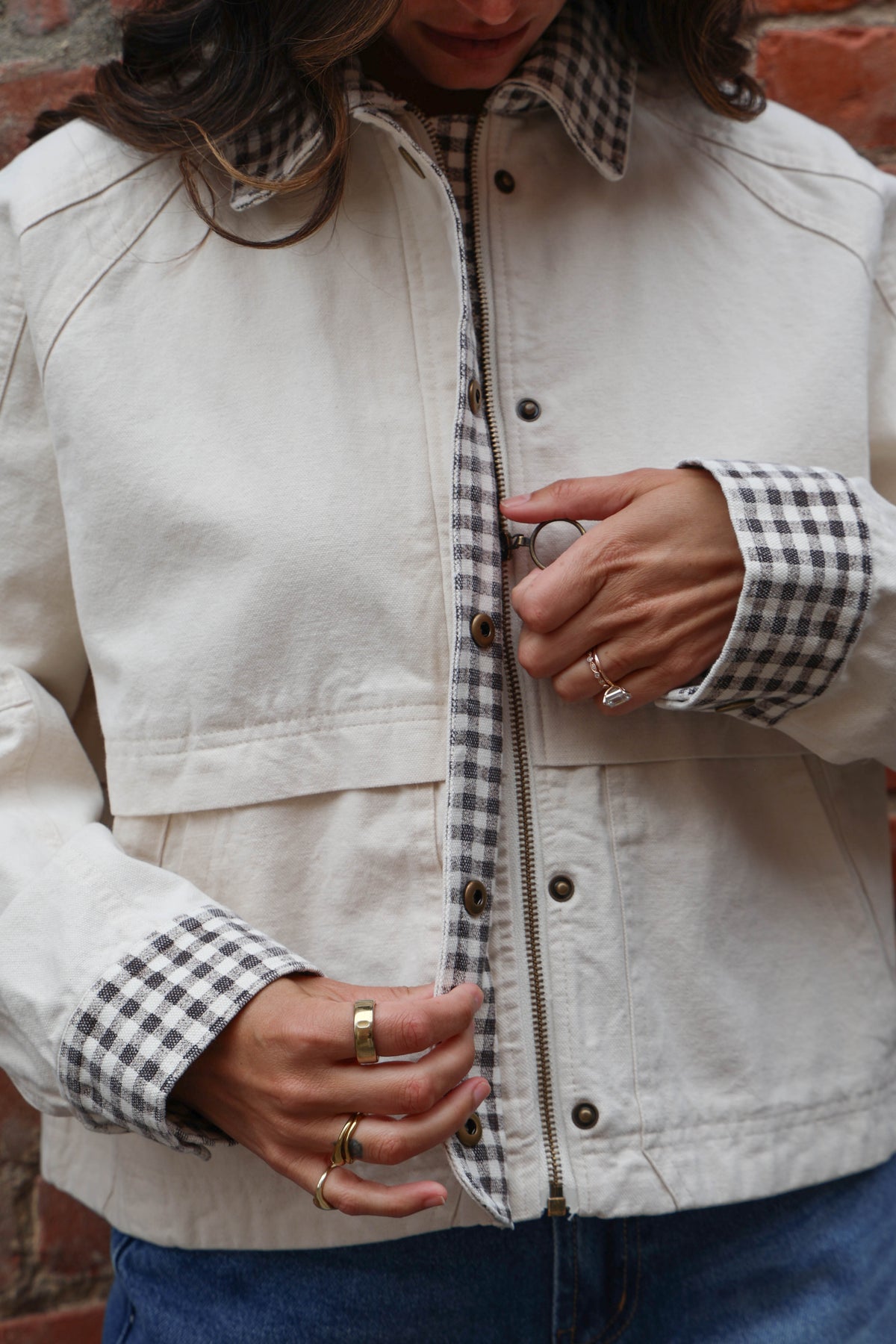 Woman in a beige jacket with gingham details and jeans against a brick wall