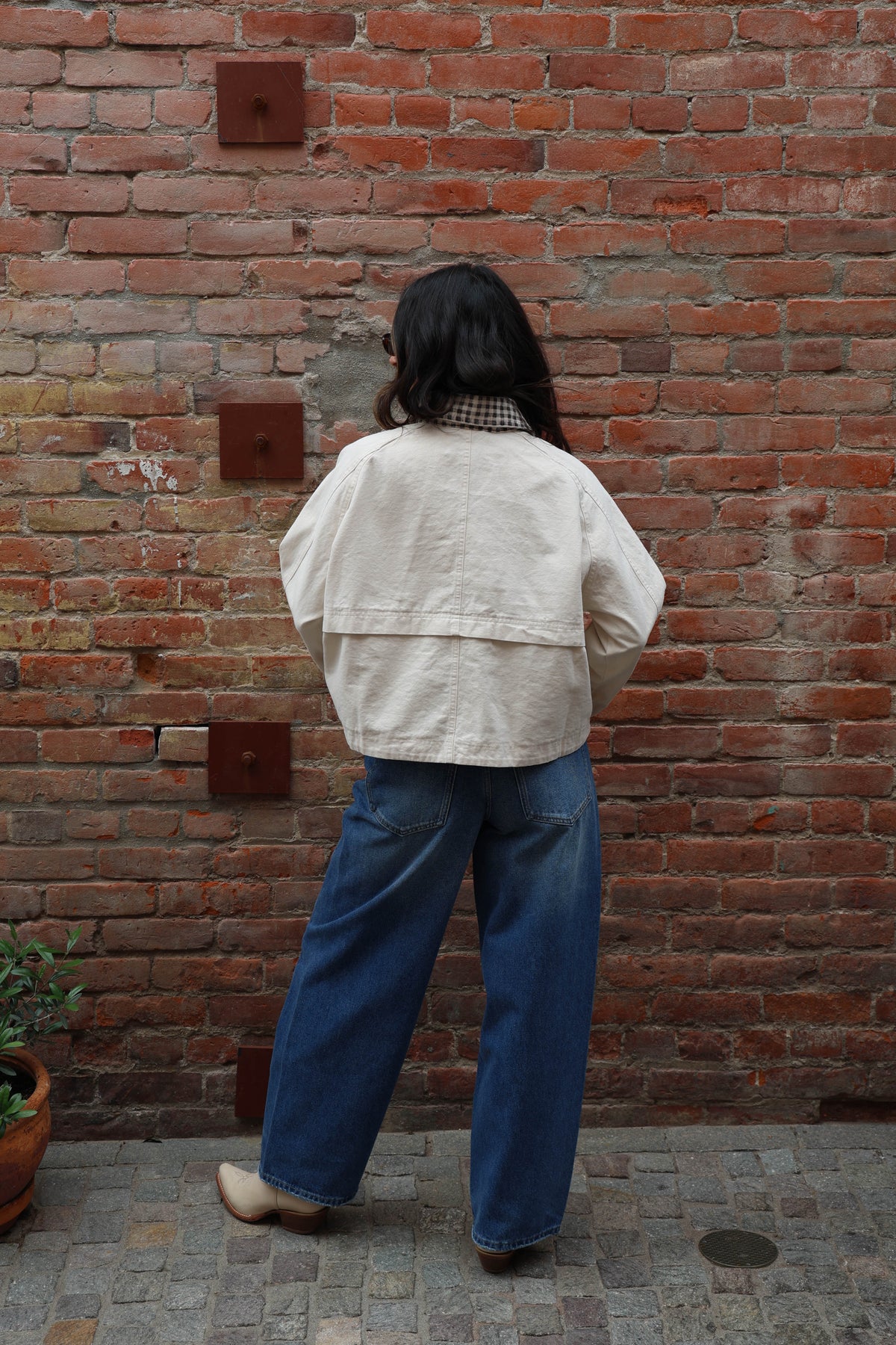 Woman in a beige jacket with gingham details and jeans against a brick wall