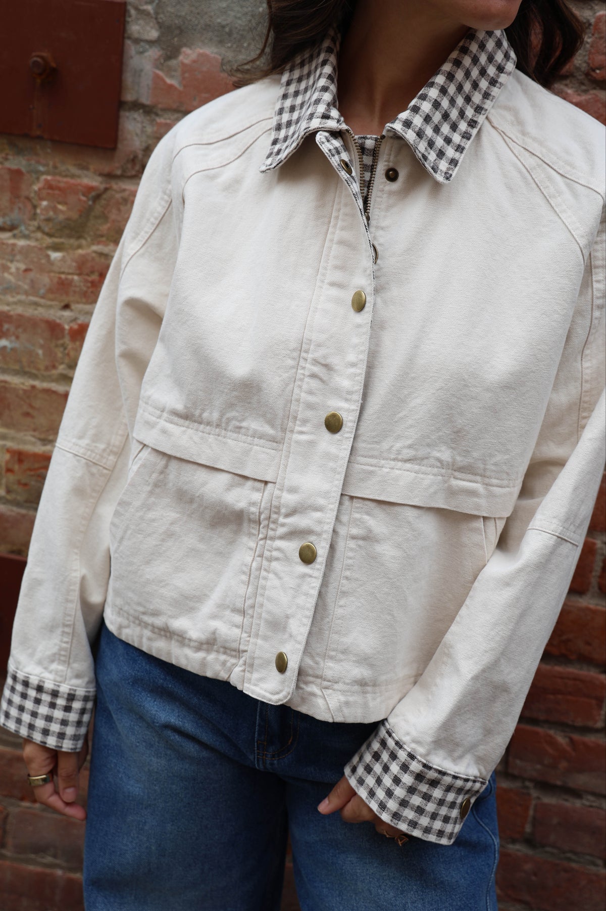 Woman in a beige jacket with gingham details and jeans against a brick wall