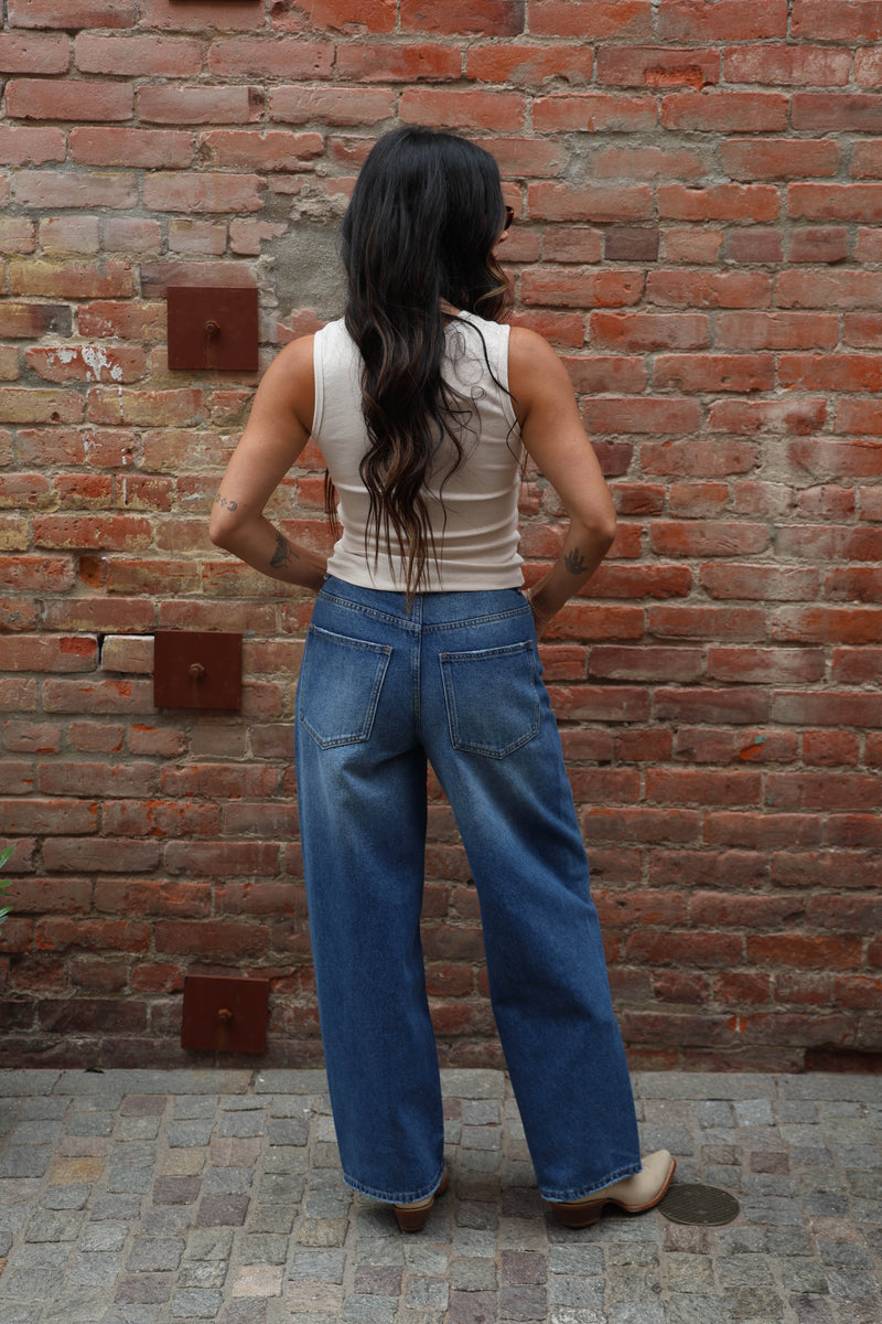 Woman standing against a brick wall wearing a white sleeveless top, blue jeans, and beige boots.
