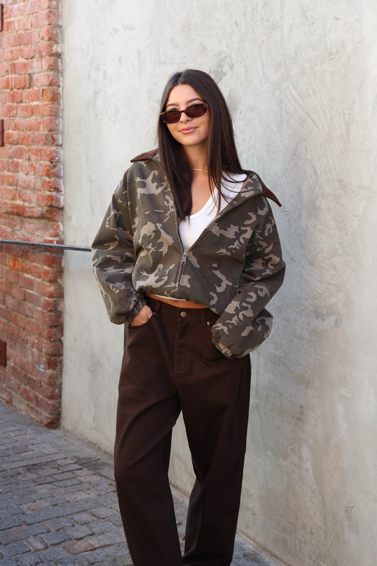 Woman wearing a camouflage jacket, white tank and brown jeans standing against a plain wall.