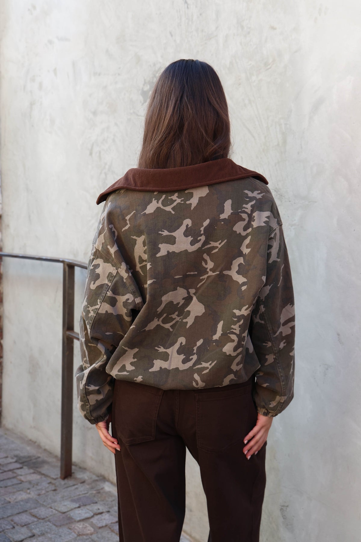 Woman wearing a camouflage jacket, white tank and brown jeans standing against a plain wall.