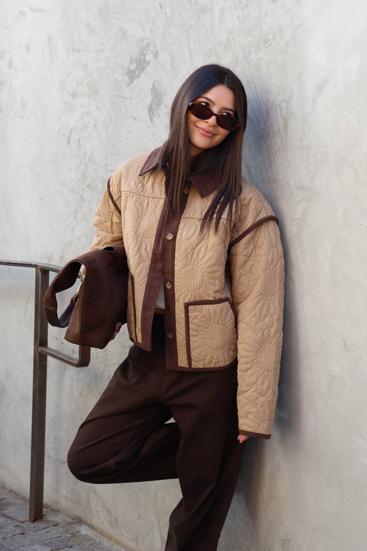 Woman wearing a brown jacket, brown pants holding a brown bad in front of a plain wall.