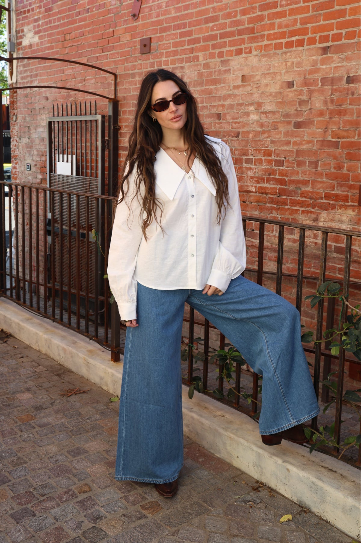 Woman in white blouse and blue jeans sitting against a brick wall.