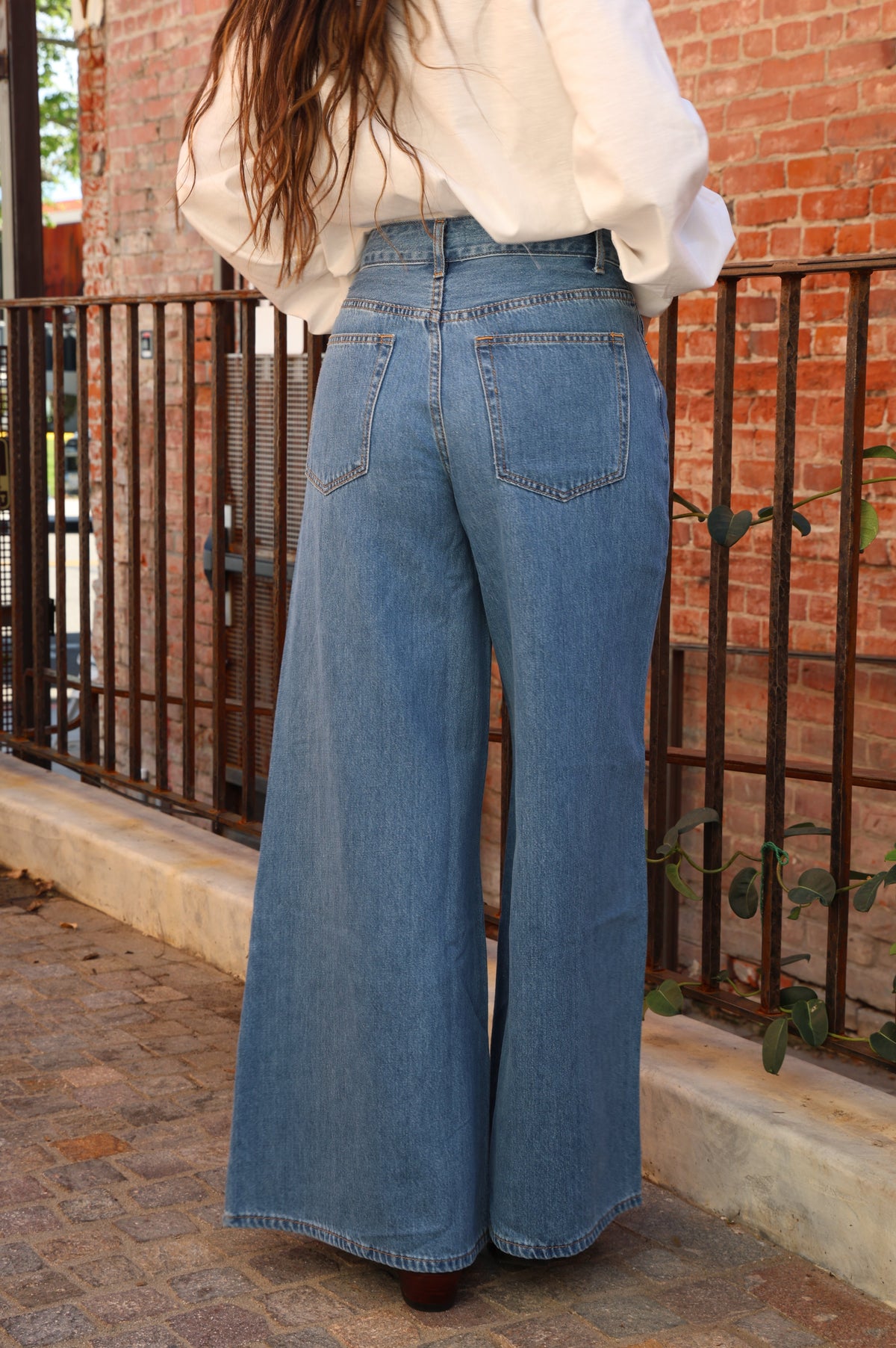 Person wearing blue jeans and a white blouse standing on a stone pavement with a brick wall in the background.