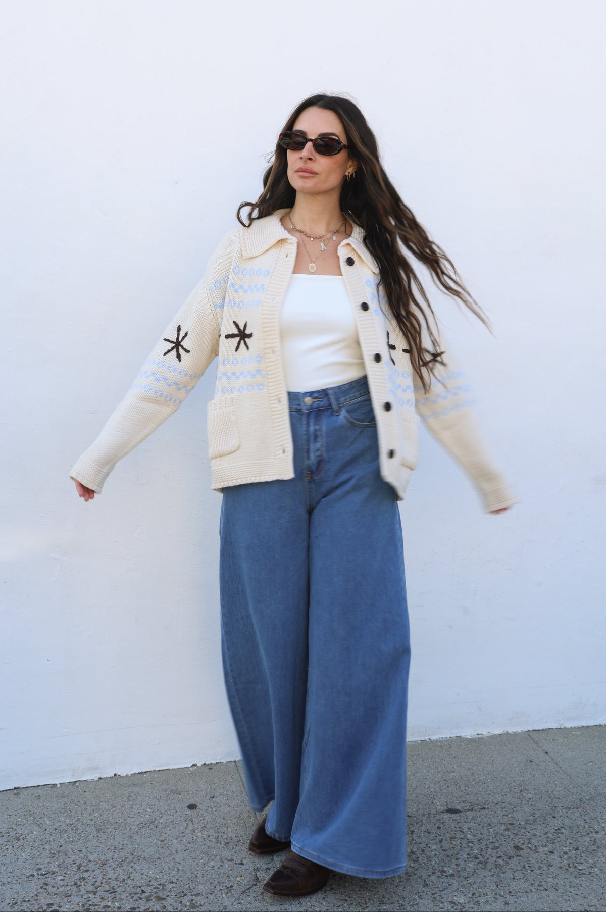 Woman wearing a beige cardigan with star patterns, white top, and blue jeans against a white wall.