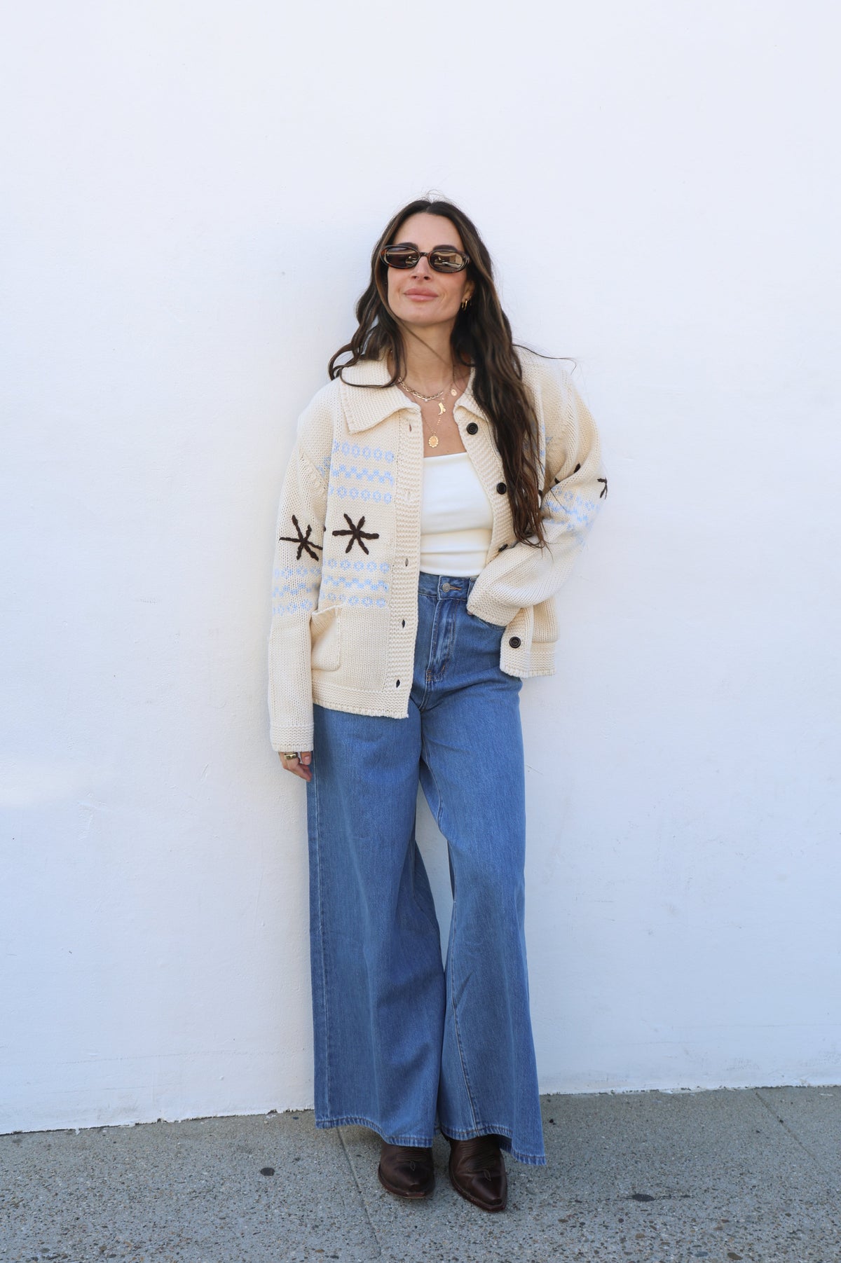 Woman wearing a beige jacket with star patterns, white top, and blue jeans against a white wall.
