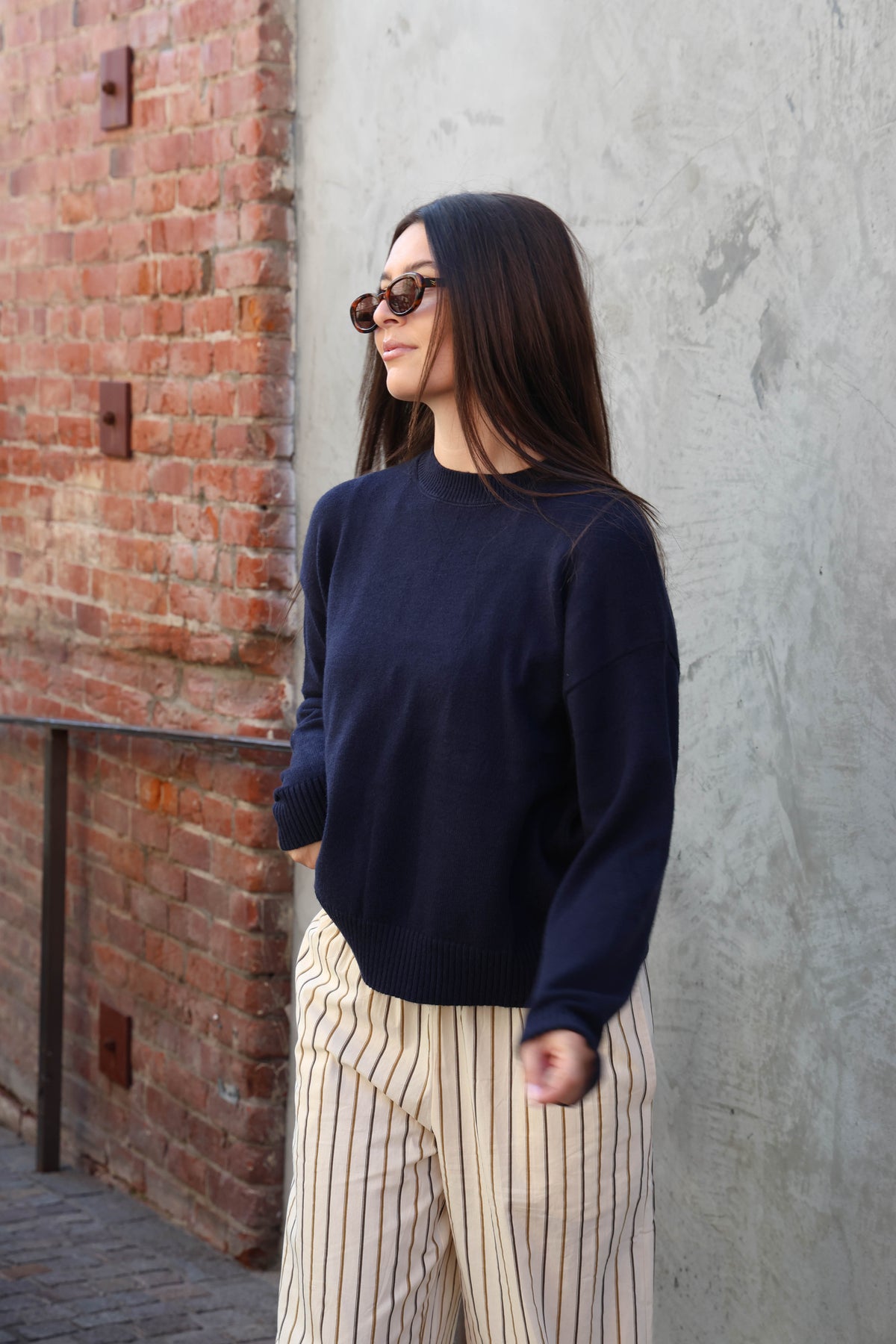 Woman wearing a sweater and striped pants standing in front of a plain wall.