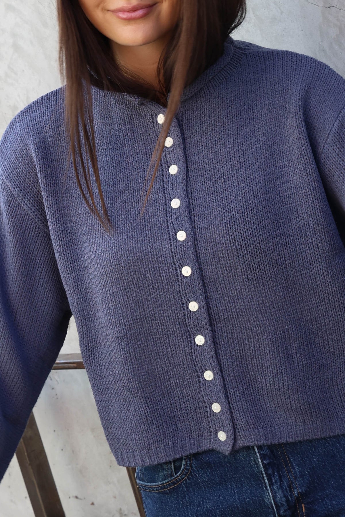 Woman wearing jeans and a blue sweater with buttons standing in front of a plain wall.