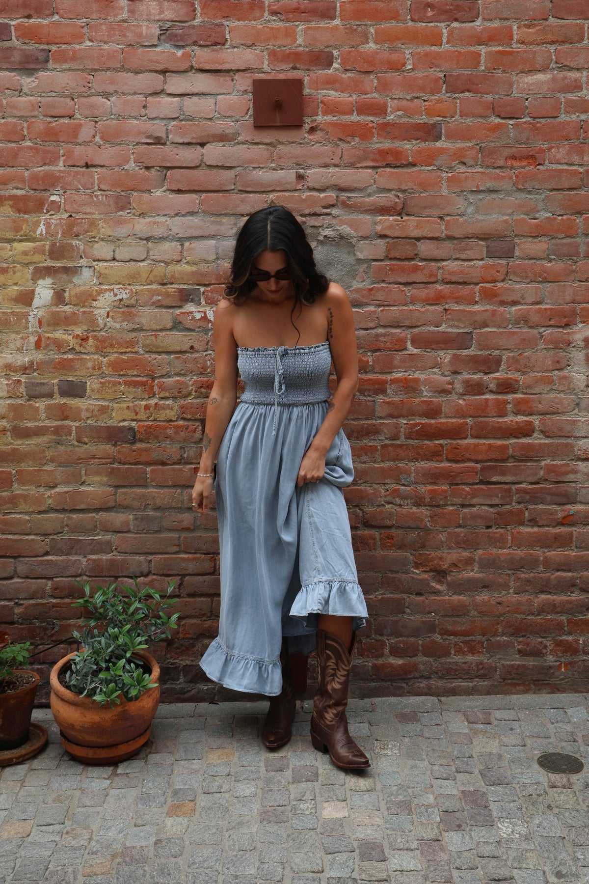 Woman in a blue dress and brown boots standing against a brick wall.