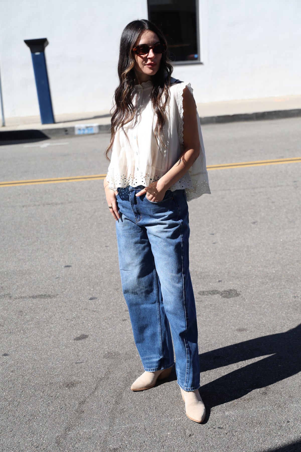 Person wearing blue jeans and a white top standing on a street with a building in the background.