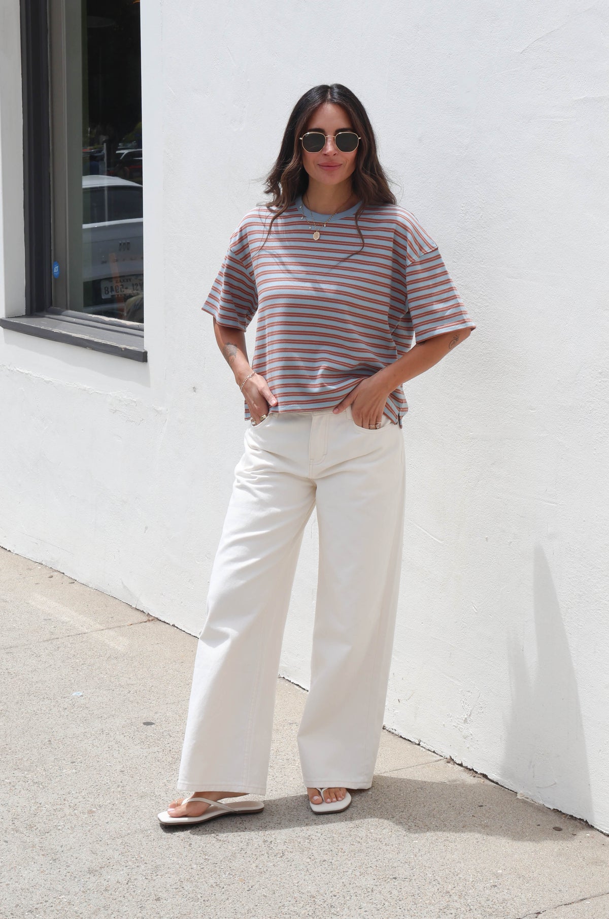 A woman stands outdoors against a white stucco wall wearing a short-sleeve striped t-shirt in muted pink, brown, and gray tones, paired with wide-leg cream pants and flat sandals.