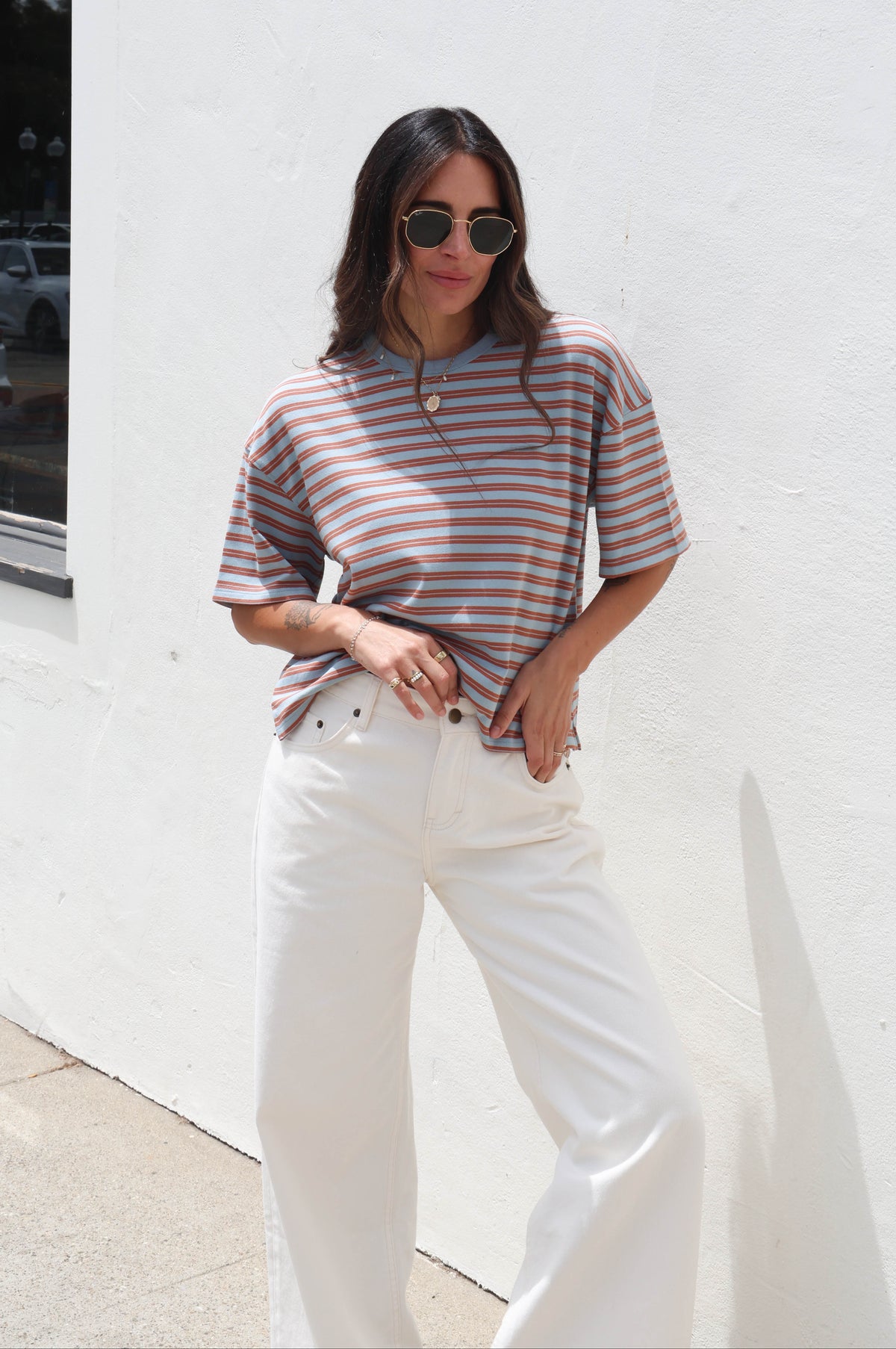 A woman wearing a short-sleeve striped knit polo shirt in muted brown and beige tones, paired with white wide-leg pants, standing against a white stucco wall.