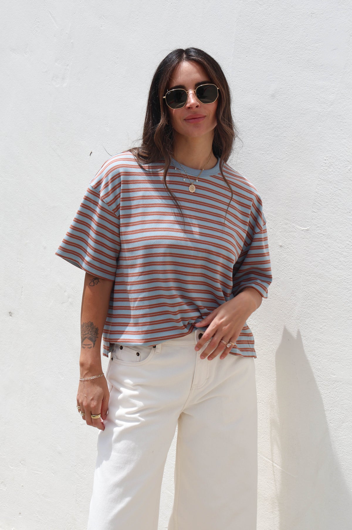 A woman wearing a short-sleeve crewneck t-shirt with thin brown and beige horizontal stripes, paired with white pants and sunglasses, standing against a white textured wall.