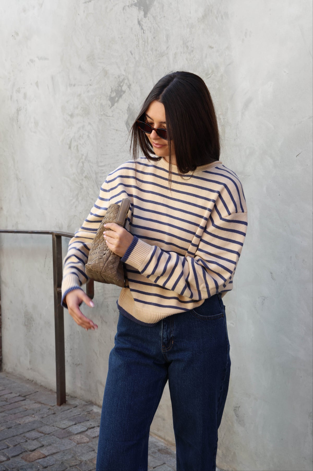 Woman wearing a cream and blue striped sweater with jeans and sunglasses standing against a plain background.