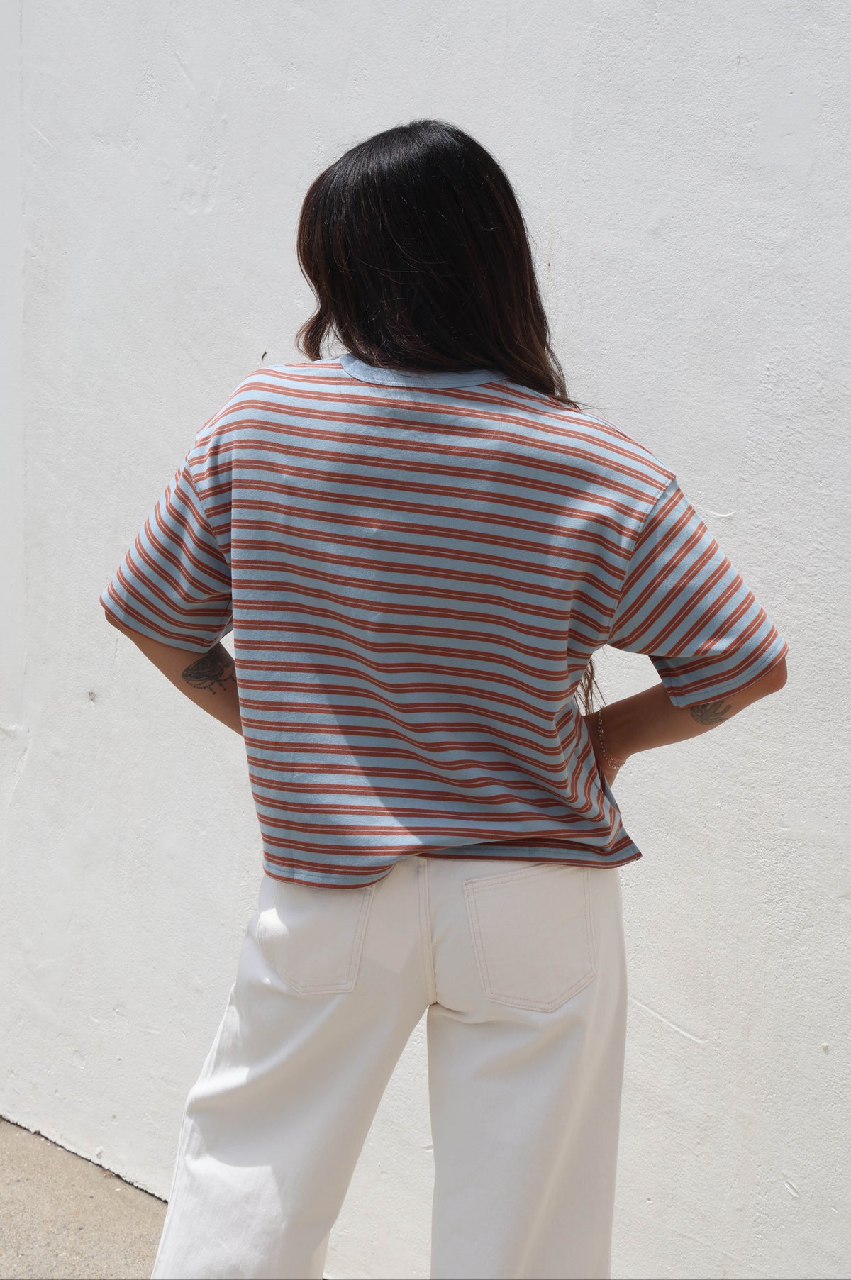 A woman viewed from behind wearing a short-sleeve striped t-shirt with orange, blue, and white horizontal stripes, paired with white pants, standing against a white stucco wall.